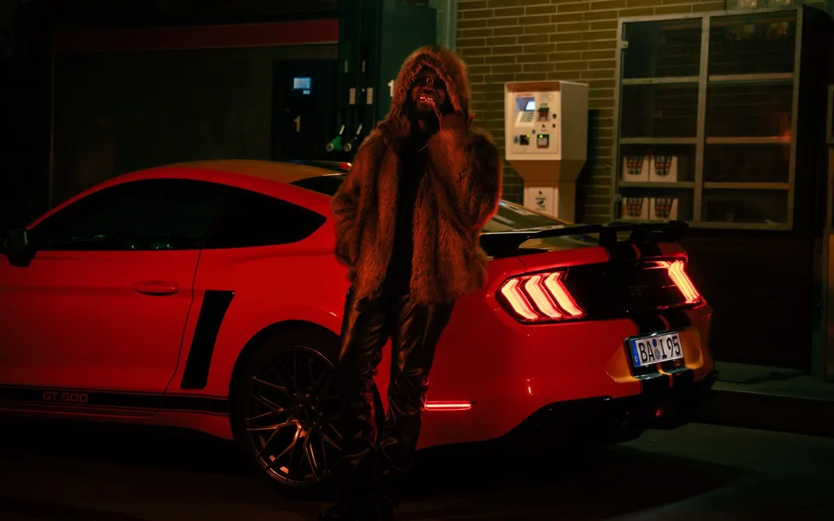 Musikvideo-Still: Artist im Fellmantel vor einem roten Ford Mustang bei Nacht